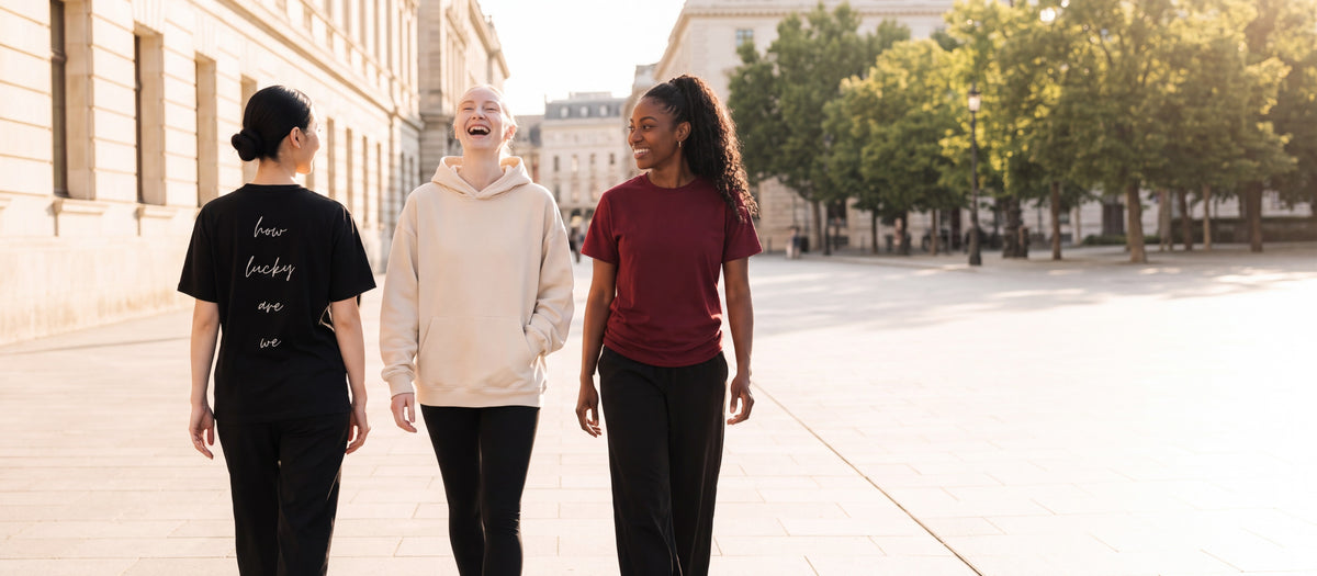 Three women walking together and laughing on a sunlit city street in golden hour light: the black How Lucky Are We tee seen from the back, the bone hoodie and a wine red tee