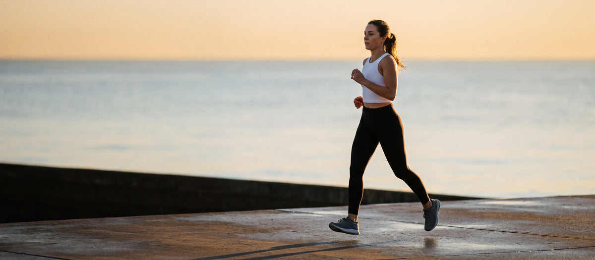 A woman running along an empty waterfront promenade at sunrise in plain black leggings and a white cropped tank