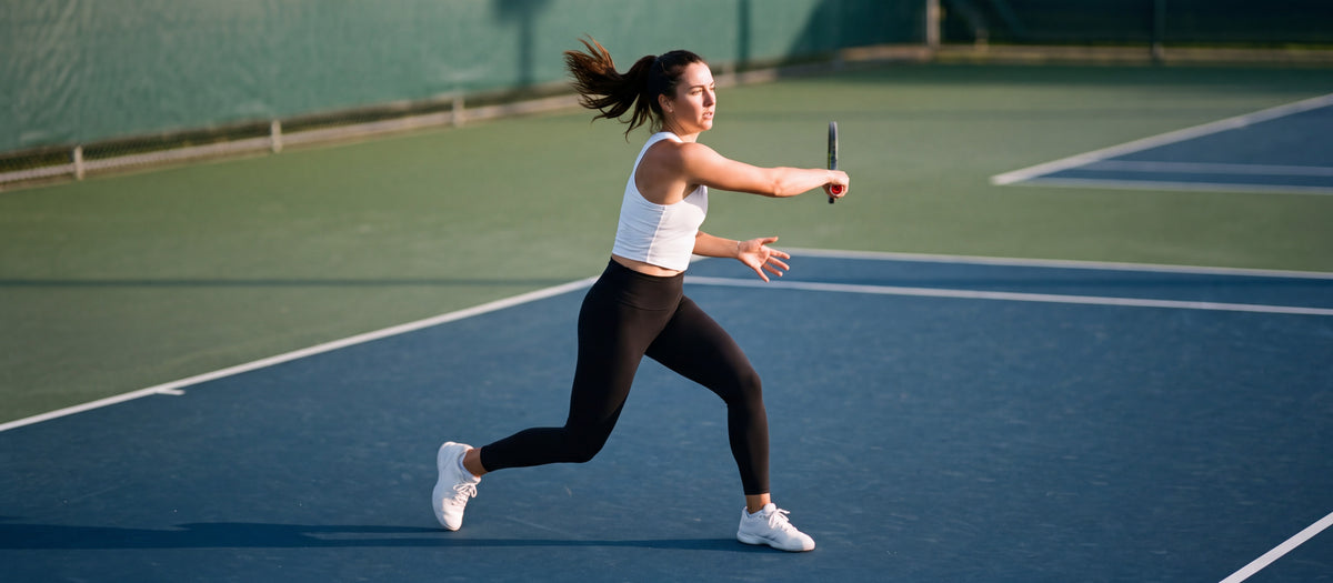 A woman mid-forehand on an outdoor tennis court at sunrise in a plain white cropped tank and black leggings